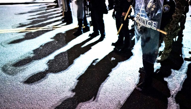 A line of police with riot shields on the street cast shadows onto the asphalt